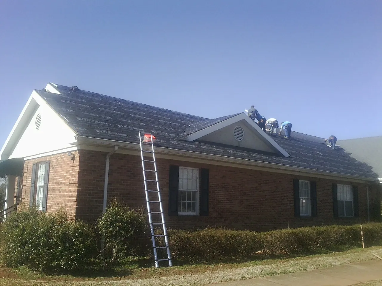 Crew tearing shingles off a brick ranch roof with a ladder set — Emergency Roof Repair work by our crew in Central